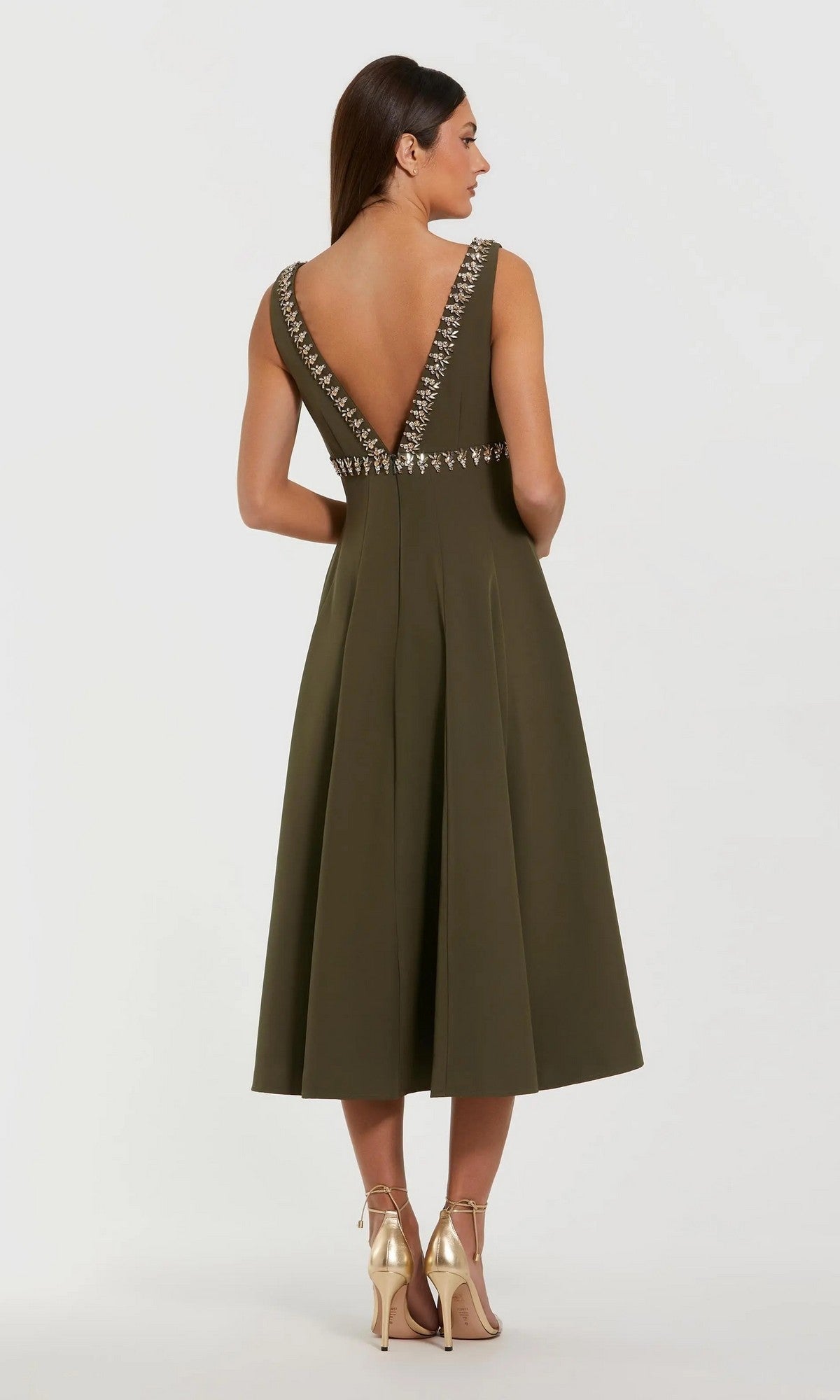 A woman in the olive green Mac Duggal 21159 midi wedding guest dress, featuring decorative trim and a V-back, pairs it with gold heels against a plain background—an elegant look for attending weddings.